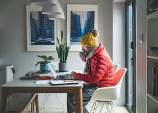 Mature man sitting working from home in a red puffer coat, scarf and wooly hat - Cost of Living Crisis
