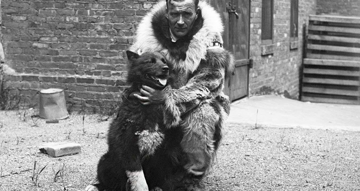 A black-and-white photo of a man dressed in warm furs crouching down next to Balto, a dog that appears to be a dark-furred husky type dog. Behind them is a brick building.