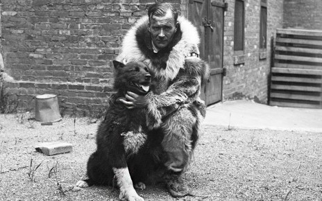 A black-and-white photo of a man dressed in warm furs crouching down next to Balto, a dog that appears to be a dark-furred husky type dog. Behind them is a brick building.