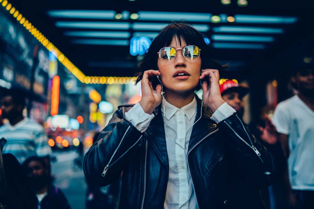 Half length portrait of trendy female millennial in electronic spectacles looking at camera during time for listening audio book and walk in metropolitan downtown, generation z in earphones; Shutterstock ID 1722211435; purchase_order: -; job: -; client: -; other: -