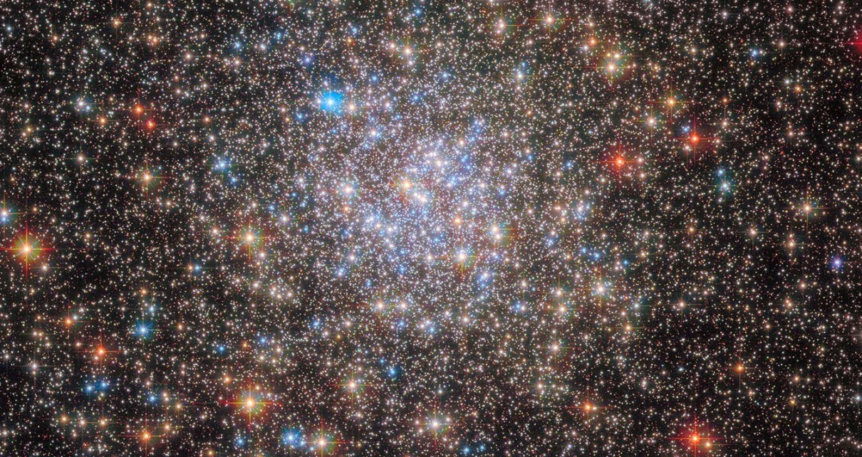 A dense collection of stars covers the view. Towards the centre the stars become even more dense in a circular region, and also more blue. Around the edges there are some redder foreground stars, and many small stars in the background.