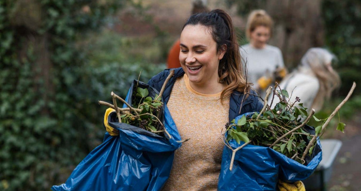 A front view of a young brunette woman carrying two bags of garden waste that is to be composted and reused. She is working on a community space garden in Hexham in the North East of England with a group of friends.
