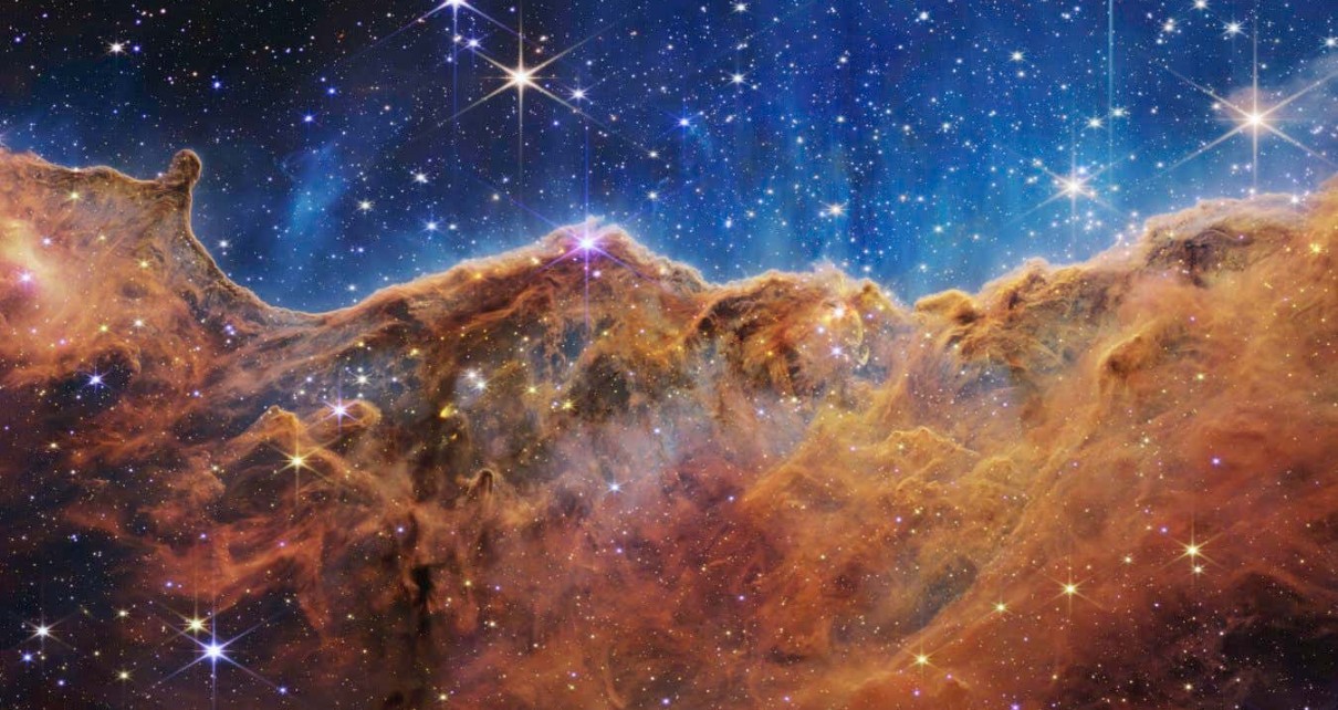 What looks much like craggy mountains on a moonlit evening is actually the edge of a nearby, young, star-forming region NGC 3324 in the Carina Nebula. Captured in infrared light by the Near-Infrared Camera (NIRCam) on NASA???s James Webb Space Telescope, this image reveals previously obscured areas of star birth. Called the Cosmic Cliffs, the region is actually the edge of a gigantic, gaseous cavity within NGC 3324, roughly 7,600 light-years away. The cavernous area has been carved from the nebula by the intense ultraviolet radiation and stellar winds from extremely massive, hot, young stars located in the center of the bubble, above the area shown in this image. The high-energy radiation from these stars is sculpting the nebula???s wall by slowly eroding it away. NIRCam ??? with its crisp resolution and unparalleled sensitivity ??? unveils hundreds of previously hidden stars, and even numerous background galaxies. Several prominent features in this image are described below. ??? The ???steam??? that appears to rise from the celestial ???mountains??? is actually hot, ionized gas and hot dust streaming away from the nebula due to intense, ultraviolet radiation. ??? Dramatic pillars rise above the glowing wall of gas, resisting the blistering ultraviolet radiation from the young stars. ??? Bubbles and cavities are being blown by the intense radiation and stellar winds of newborn stars. ??? Protostellar jets and outflows, which appear in gold, shoot from dust-enshrouded, nascent stars. ??? A ???blow-out??? erupts at the top-center of the ridge, spewing gas and dust into the interstellar medium. ??? An unusual ???arch??? appears, looking like a bent-over cylinder. This period of very early star formation is difficult to capture because, for an individual star, it lasts only about 50,000 to 100,000 years ??? but Webb???s extreme sensitivity and exquisite spatial resolution have chronicled this rare event. Located roughly 7,600 light-years away, NGC 3324 was first cat