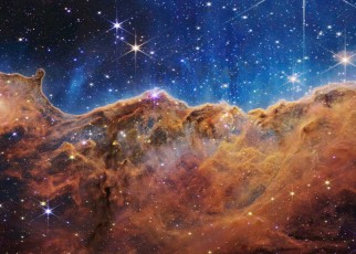 What looks much like craggy mountains on a moonlit evening is actually the edge of a nearby, young, star-forming region NGC 3324 in the Carina Nebula. Captured in infrared light by the Near-Infrared Camera (NIRCam) on NASA???s James Webb Space Telescope, this image reveals previously obscured areas of star birth. Called the Cosmic Cliffs, the region is actually the edge of a gigantic, gaseous cavity within NGC 3324, roughly 7,600 light-years away. The cavernous area has been carved from the nebula by the intense ultraviolet radiation and stellar winds from extremely massive, hot, young stars located in the center of the bubble, above the area shown in this image. The high-energy radiation from these stars is sculpting the nebula???s wall by slowly eroding it away. NIRCam ??? with its crisp resolution and unparalleled sensitivity ??? unveils hundreds of previously hidden stars, and even numerous background galaxies. Several prominent features in this image are described below. ??? The ???steam??? that appears to rise from the celestial ???mountains??? is actually hot, ionized gas and hot dust streaming away from the nebula due to intense, ultraviolet radiation. ??? Dramatic pillars rise above the glowing wall of gas, resisting the blistering ultraviolet radiation from the young stars. ??? Bubbles and cavities are being blown by the intense radiation and stellar winds of newborn stars. ??? Protostellar jets and outflows, which appear in gold, shoot from dust-enshrouded, nascent stars. ??? A ???blow-out??? erupts at the top-center of the ridge, spewing gas and dust into the interstellar medium. ??? An unusual ???arch??? appears, looking like a bent-over cylinder. This period of very early star formation is difficult to capture because, for an individual star, it lasts only about 50,000 to 100,000 years ??? but Webb???s extreme sensitivity and exquisite spatial resolution have chronicled this rare event. Located roughly 7,600 light-years away, NGC 3324 was first cat