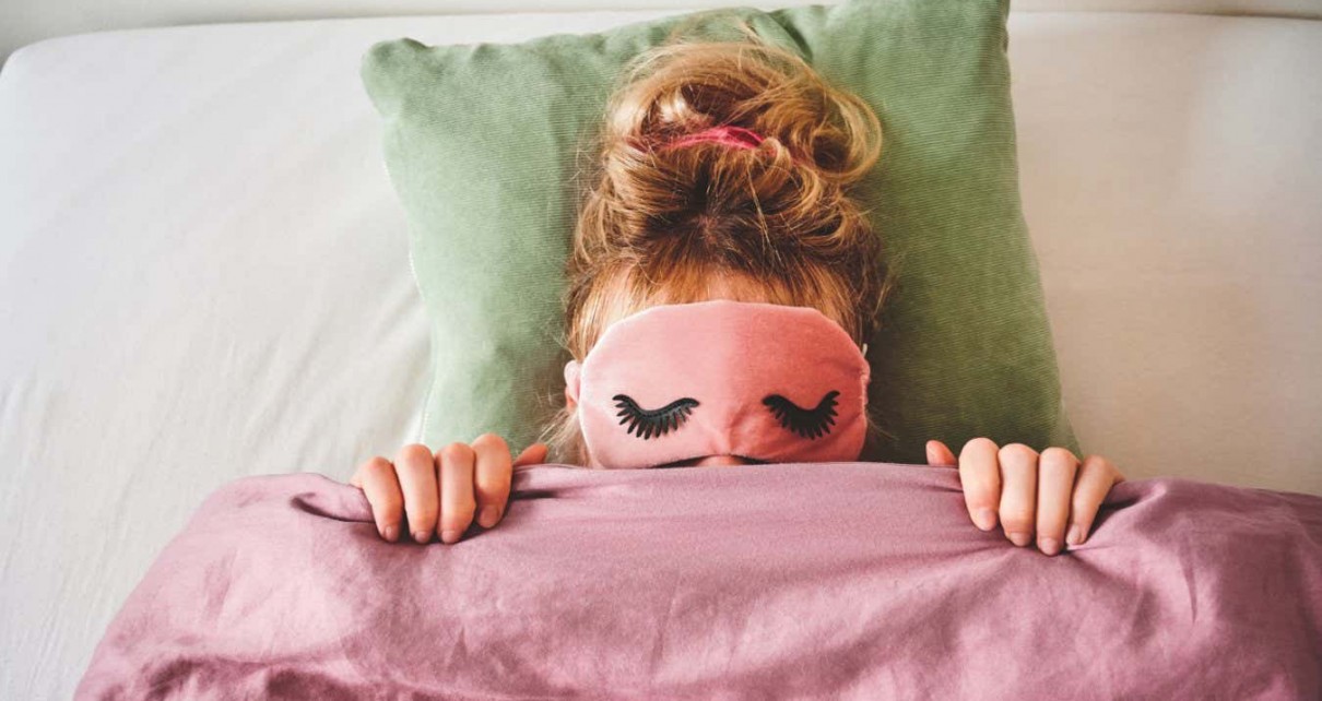 A woman in bed with a sleep mask on and covering her face with her duvet