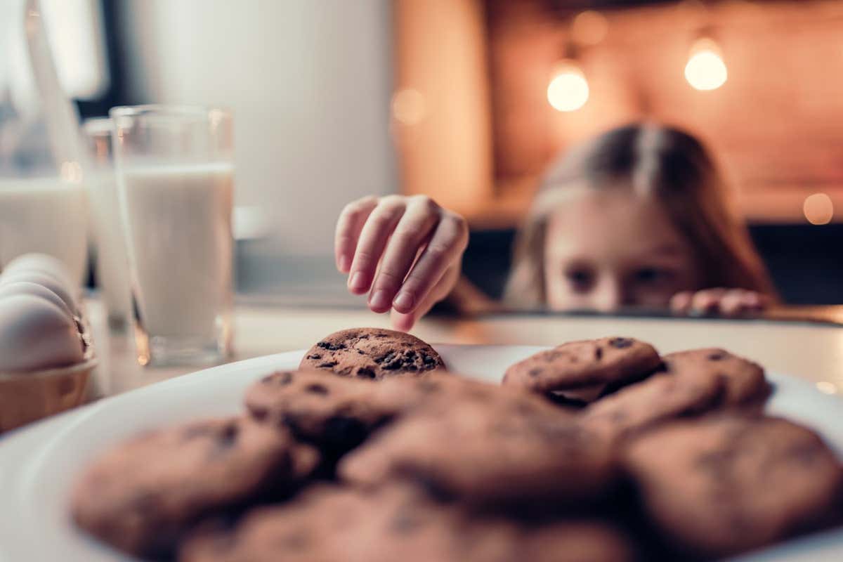Little cute girl on kitchen is taking chocolate cookie from table.