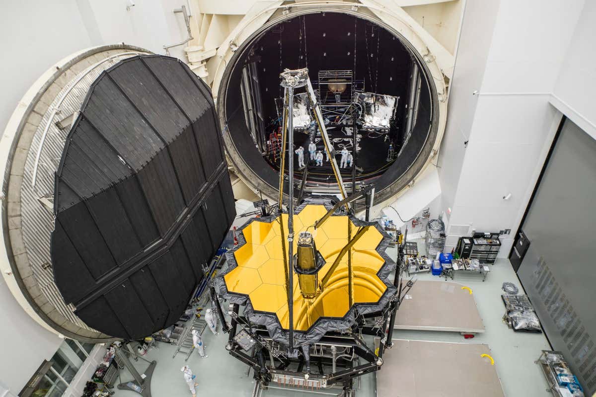A fully deployed OTIS, with the secondary mirror extended over the primary mirror, is removed from Chamber A after a successful thermal-vacuum test in 2017. Seen still in the chamber is a sunshield simulator, called the L5 Simulator, representing the fifth and innermost layer of the sunshield. The telescope would be shipped to the Northrop Grumman facility in California to be integrated with the real sunshield. I was suspended on a lift for this shot. Page 119