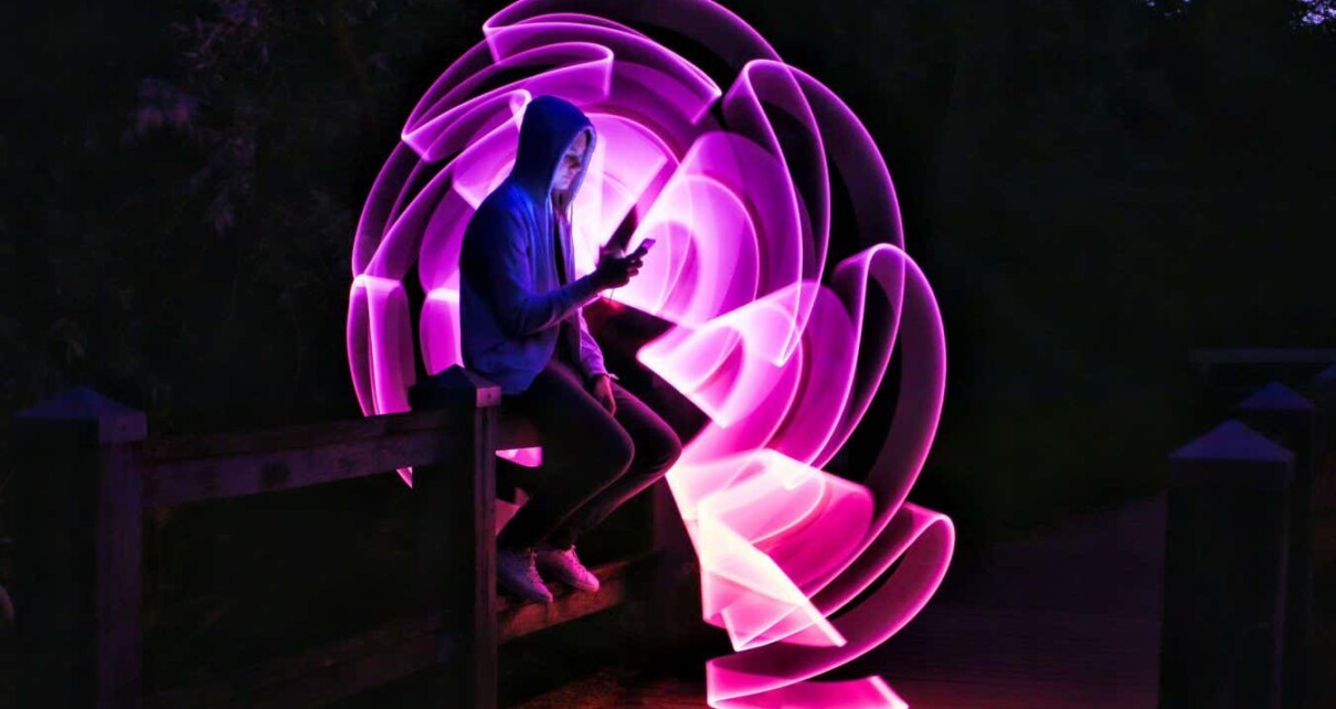 2C72973 Boy sitting on a bridge with his smartphone in his hand. Violet light effect with a light saber in lightpainting.