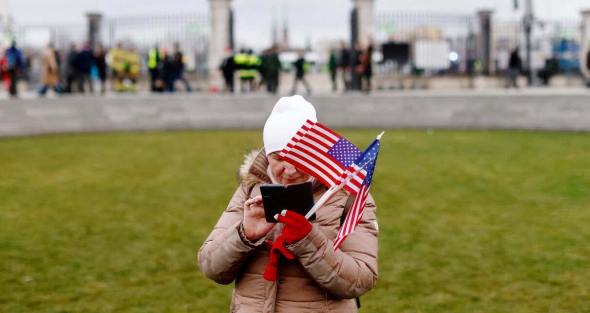A woman holding several small flags of the United States checks her mobile phone while waiting with other onlookers prior to the arrival of the US President in Warsaw on February 21, 2023. - US President Biden is due to deliver a speech in Warsaw later on February 21 at Royal Warsaw Castle Gardens. (Photo by Wojtek Radwanski / AFP) (Photo by WOJTEK RADWANSKI/AFP via Getty Images)