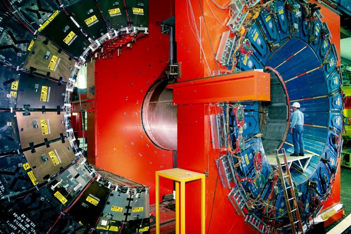 CDF particle detector at the Fermi National Accelerator Laboratory (Fermilab) near Chicago, USA. The CDF (Collider Detector Facility) records subatomic particles created in high-energy proton-antiproton collisions in the Tevatron coll- ider. It co-discovered the top quark in 1994. Here the detector is partly disassembled between operation runs. The black segments (left) are part of the outer muon chambers. The red areas are part of the magnet which bends charged particles travelling out from the collision point. The blue segments (right) are part of the hadron calorimeter. When in use these parts are closed tightly together. Photographed in 1998. The physicist is ROB ROSER.